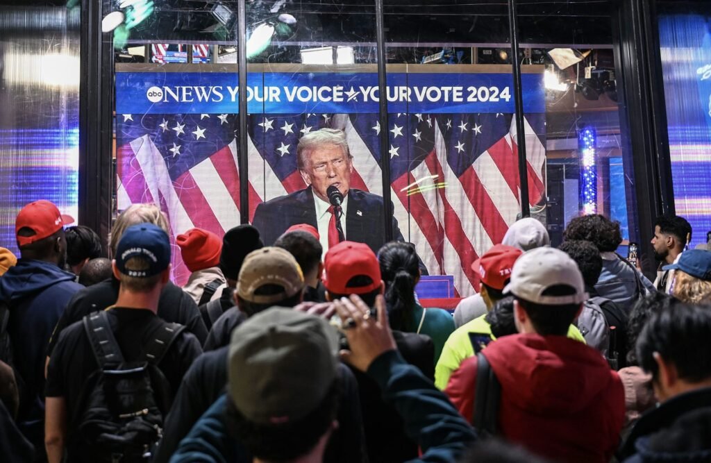 Several people wearing baseball hats watch a man speak on TV.